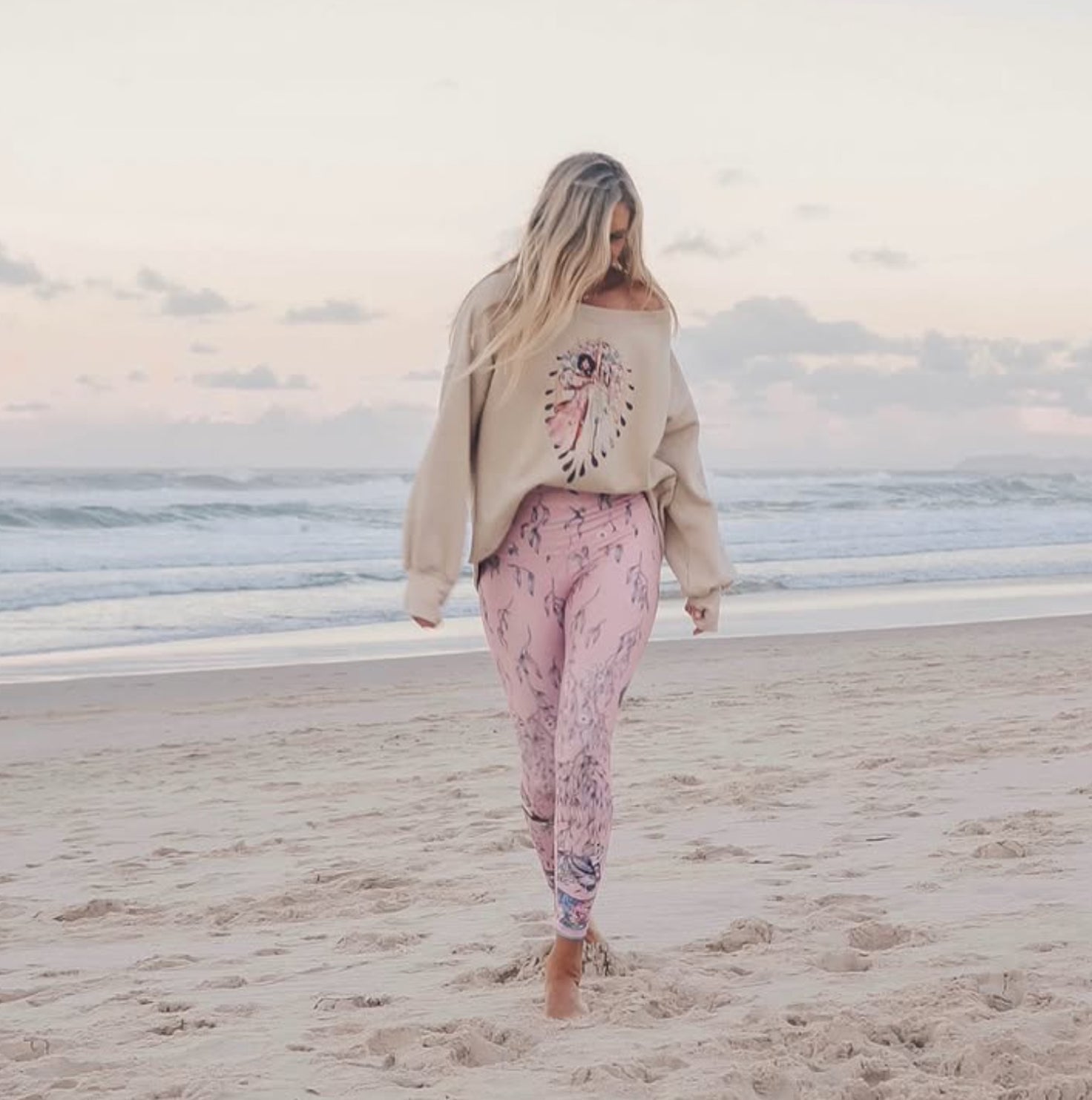 Woman walking on the beach wearing pastel pink leggings and a beige sweatshirt with an artistic print — conscious clothing by Studio Hannah Chloe.