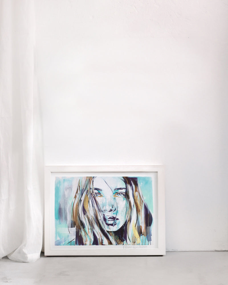 Bohemian portrait in turquoise of a woman showing a subtle strength. In a white frame in a plain room.